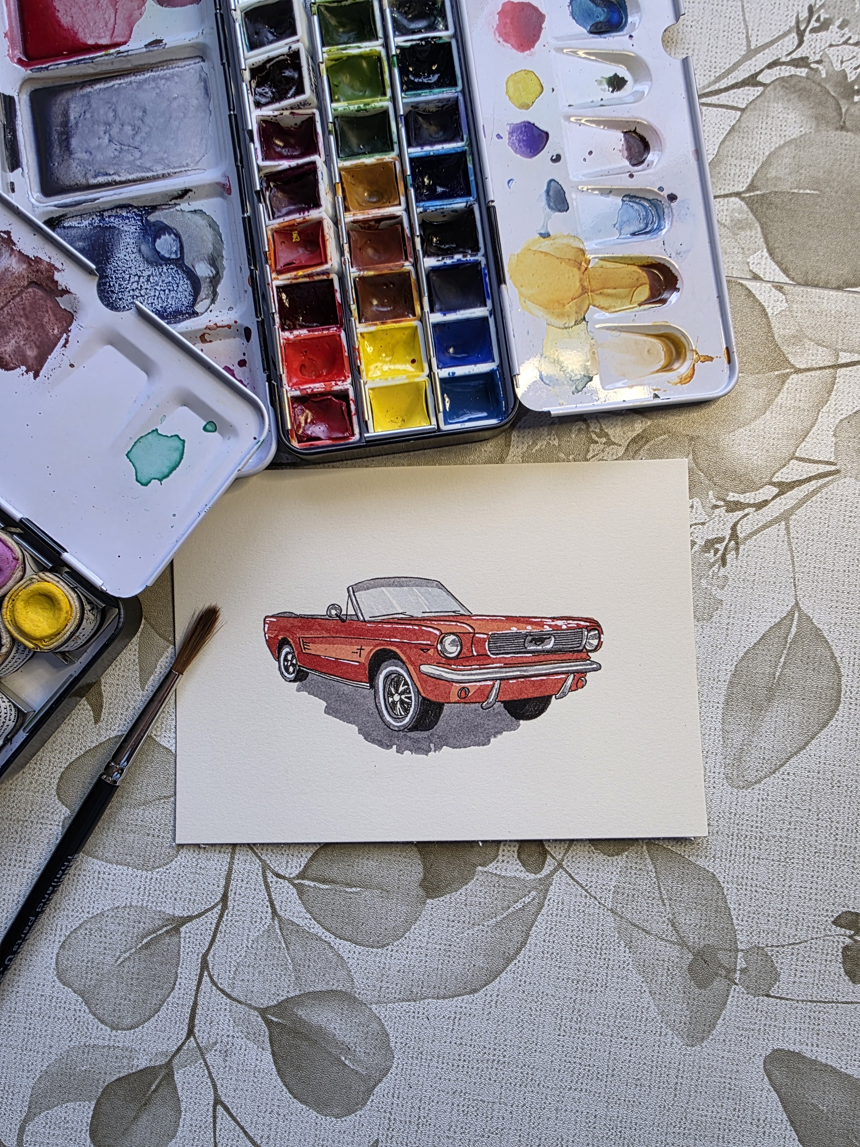 A watercolor painting of a vintage red convertible car, accompanied by a paintbrush and paint palette.